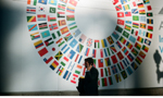 Homem vestindo terno preto, visto de perfil, falando ao celular, em frente a uma parede branca onde as bandeiras de diversos países estão dispostas em um círculo, deixando um espaço vazio no meio.