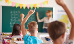 Sala de aula com três crianças de costas, erguendo a mão, como se procurassem responder a alguma pergunta da professora, que está à frente da sala, apontando para a lousa.
