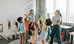 Fotografia de crianças brincando com bexigas e bolhas de sabão, supervisionadas por duas adultas, em uma sala ampla com decoração infantil e alguns tapetes dispostos no chão.