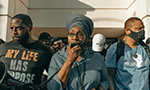 Fotografia de um grupo de pessoas negras participando de um protesto; uma mulher com um turbante, no centro, fala em um microfone, ladeada por dois homens, um usando uma máscara preta e o outro com uma camisa com as inscrições “My life has a purpose” (“Minha vida tem um propósito”, em português).