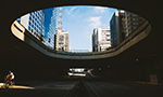 Fotografia de uma pessoa pedalando uma bicicleta dentro de um túnel. O túnel tem uma abertura circular no topo, permitindo a visão parcial dos edifícios da rua acima e do céu azul