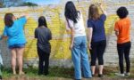 A imagem mostra cinco jovens usando roupas despojadas pintado um muro branco com tinta amarela. Usando pincéis, trabalham em equipe, dando cor ao espaço. A cena transmite colaboração e criatividade, em um ambiente urbano, iluminado pela luz do dia.